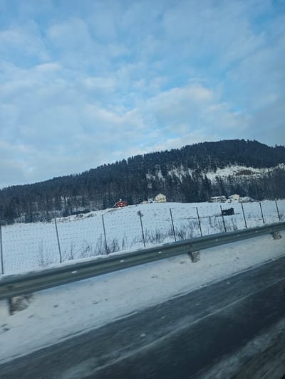 The image displays a winter landscape in Brumunddal, Norway, under an overcast sky. In the foreground, a metallic grey guardrail extends horizontally across the lower third of the frame, angled slightly upwards to the right. Below the guardrail, a dark, wet road surface is visible. Behind the guardrail, the ground is covered in snow, forming a gentle slope upwards. A light-colored fence, constructed with vertical posts and multiple horizontal wires, traverses this snow-covered field. Sparse, dark, bare vegetation protrudes from the snow within the fenced area. Two distinct structures are visible on the upward slope: a red-painted house situated on the left, and a larger, lighter-colored (possibly cream or light yellow) house located to its right. Further to the right and partially obscured behind the fence, a dark, irregularly shaped object is present. The background is dominated by a densely forested hill or mountain, covered in dark evergreen trees, with visible patches of snow on its slopes. The sky above features light blue areas interspersed with greyish-white clouds. No individuals, explicit actions, or direct interactions are depicted in the scene.
