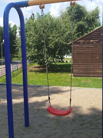 A red plastic swing seat hangs by rope from a metal frame in a sandy playground. The swing is empty. In the background, a wooden building with a sloped roof stands behind a dense green tree line. A well-maintained lawn is visible beyond the sand. The sky above is partly cloudy. The scene appears to be in an outdoor public space.