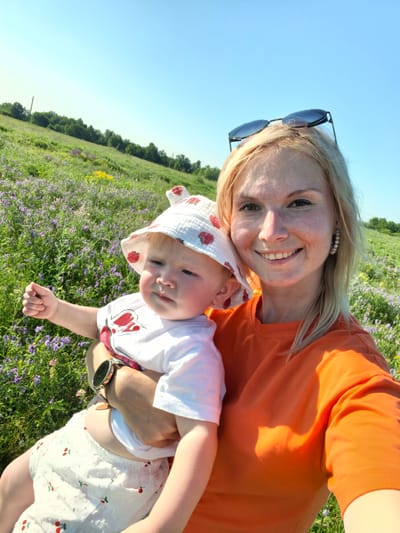 A woman and a child are in a field of purple flowers under a clear blue sky. The woman, on the right, has blonde hair and is wearing an orange t-shirt and sunglasses on her head. She is smiling at the camera. She is holding a child, on the left, who is wearing a white hat with red strawberries and a white t-shirt with a red design. The child is looking to the left and has their hand raised. The field is covered in purple and yellow flowers, with trees in the background. The location is a field outside of Karaiganovo, Russia.