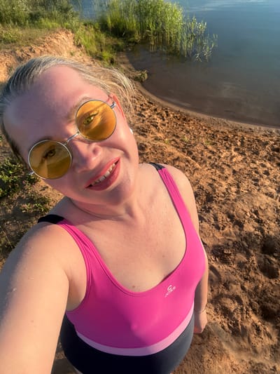 This is a selfie taken by a woman lying on a sandy shore next to a body of water. She is wearing a pink swimsuit with black accents and yellow-tinted round sunglasses. Her mouth is slightly open, revealing her teeth, and she has pink lipstick on. She appears to be looking up at the camera. The background shows sand, some green vegetation, and a large area of water. The lighting suggests it is daytime, possibly late afternoon or early evening given the warm tones. The overall impression is one of relaxation and enjoying a sunny day outdoors. The location is identified as Sapunov Bor, Russia. There is a visible logo on her swimsuit, which reads "JOS".