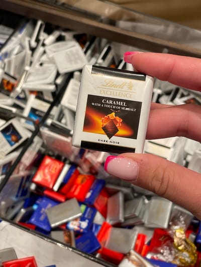 In Marratxí, Spain, a close-up image depicts a human hand holding a single, individually wrapped Lindt Excellence "Caramel with a Touch of Sea Salt Dark-Noir" chocolate square. The chocolate wrapper is predominantly white, featuring the Lindt logo and product description, with a graphic of caramel flowing over a dark chocolate piece. The hand, likely female, displays a manicured index finger and thumb, both with pink French tip nail polish. The chocolate is held above a large, deep bin or container, which is filled with an assortment of various individually wrapped chocolates. These background chocolates are largely out of focus but reveal a mix of white, red, blue, and silver wrappers, suggesting multiple flavors or brands, with many appearing to be additional Lindt squares.