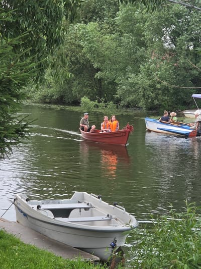 In this vertically oriented image, several people are enjoying a day on the water. The scene appears to be a calm river or lake, with lush green trees lining the banks and a bright, overcast sky suggesting either early morning or late afternoon.
The primary focus is on a brown rowboat in the center, carrying three individuals wearing orange life vests. One person is rowing, while the other two are seated. To the left, another rowboat with blue and white accents is visible, occupied by at least two people. A white boat is docked on the right side of the frame, grounded on a concrete or wooden edge next to a patch of vibrant green grass.
The water reflects the surrounding greenery and the sky, creating a tranquil atmosphere. The overall mood is relaxed and recreational, hinting at a leisurely outing. There is no visible text in the image. The location context suggests this scene is set in Pereslavl-Zalessky, Russia.
