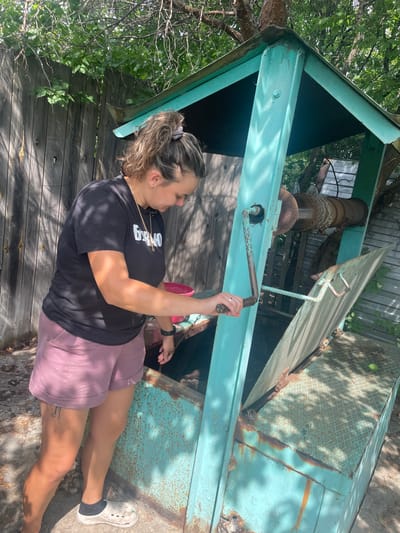 In the village of Raslovka 1-Ya, Russia, a woman with her hair tied in a bun, wearing a black t-shirt and pink shorts, stands next to a turquoise metal well structure. She is holding a red bucket and appears to be operating a crank mechanism on the well. The well is equipped with a chain and pulley system, and the opening is covered by a metal lid, partially ajar. The surrounding environment includes a wooden fence, trees, and greenery, suggesting an outdoor, possibly rural setting. The overall scene depicts a common task of drawing water from a well.