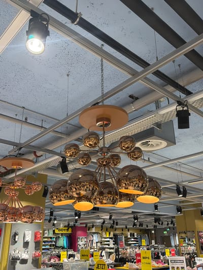 The image depicts a view of a store interior, likely in Spreitenbach, Switzerland, given the German signage. The perspective is looking upwards towards a ceiling with a complex network of metal beams, pipes, and fixtures. Two modern chandeliers with multiple reflective, golden spherical bulbs are prominently featured, one near the center and another towards the lower part of the frame. The lighting is bright, illuminating a retail space in the background where clothing displays and promotional signs for "BOYS" and "GIRLS" sections are visible. Clothing items are neatly arranged on shelves and racks. The environment is commercial, suggesting a retail store, potentially a clothing store in a shopping center or mall.