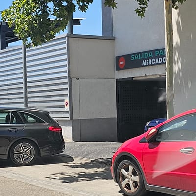 The image depicts an outdoor scene near a parking garage entrance in Cuarte de Huerva, Spain. A black station wagon is parked on the left side of the frame, with its rear visible. In the foreground, a red hatchback is partially visible, with its front right wheel and side of the car in view. The parking garage entrance is a dark, metal grate structure, and above it, a sign reads "SALIDA PARKING MERCADO" in green lettering. A small, red circular sign with a white bar is also visible on the wall to the left of the entrance, possibly indicating an exit or restricted area. A grey metal corrugated wall is visible behind the black station wagon, and a tree with green leaves casts shadows on the ground. The sky is blue and clear. There are no people clearly visible in the image.