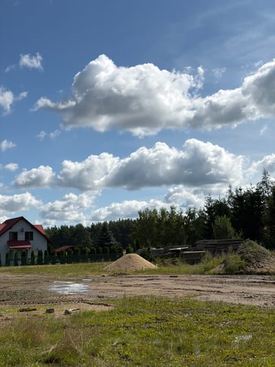 This image depicts a partially constructed residential area in Skrzeszewo, Poland. In the foreground, a muddy field is dotted with puddles of water and patches of green grass. Several construction materials, including a pile of sand and a stack of lumber, are visible. A white house with a red roof stands in the mid-ground, bordered by a green fence and manicured hedges. Behind the house, a dense forest of trees forms the background. The sky above is a clear blue, adorned with numerous fluffy white clouds. The scene suggests ongoing construction and development in a semi-rural setting.