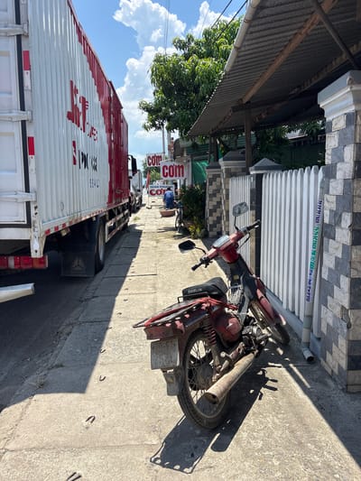 In a narrow urban alleyway, a large white truck with red lettering is parked on the left, its side taking up a significant portion of the frame. To the right of the truck, a weathered red motorcycle is parked parallel to a white picket fence. The fence is constructed of vertical wooden slats and is attached to stone pillars. Behind the fence, a corrugated metal roof extends over a walkway. Two signs with the word "com" are visible on the right side of the alley, near a person in blue attire. The ground of the alley is paved with concrete, with a few U-shaped metal fixtures embedded in it. The scene is illuminated by bright sunlight, casting distinct shadows.
