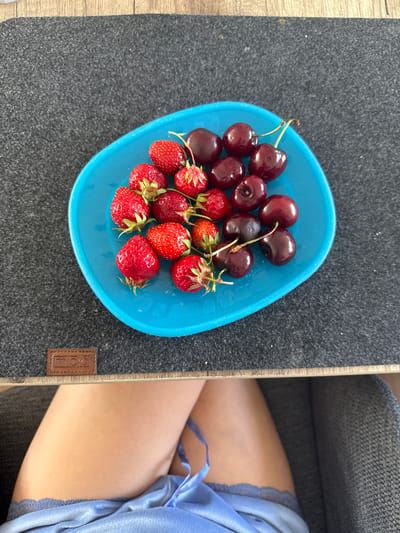 This image depicts an indoor scene in Bruchköbel, Germany, viewed from a slightly elevated perspective. A light blue, shallow, oval-shaped bowl, positioned centrally on a dark gray felt mat, contains a mixture of approximately 10-12 ripe red strawberries with green caps and stems, and 10-12 dark red cherries, some with green/yellow stems attached. The felt mat rests on a light wooden surface. A small, brown rectangular tag, embossed with the word "Miqio", is visible on the mat in the lower-left quadrant. Below the table level, the bare legs of a person are visible, crossed at the knees, wearing light blue undergarments with a lace trim. A portion of a dark gray textured fabric chair is present to the right of the person's legs. The scene is brightly and evenly lit.