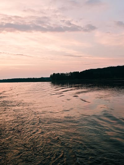 A body of water reflects the pink and orange hues of a sunset sky. A dark line of trees forms the horizon. The water's surface is rippled, creating patterns of light and shadow. A small, dark object, possibly a buoy or a distant person, is visible in the water to the left. The overall impression is of a tranquil, natural scene at dusk. The location is the Volga River near Nakhval'skoe, Russia.