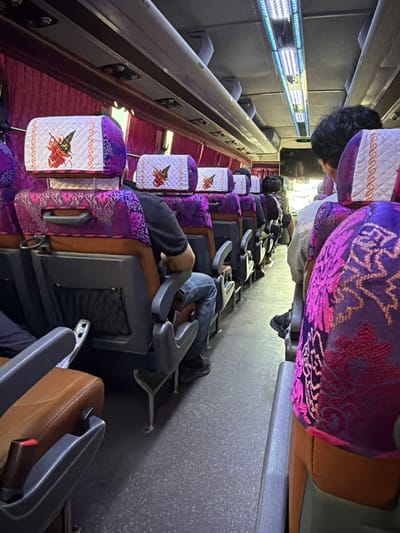 The image shows the interior of a bus with rows of purple and patterned seats. Several passengers are seated, facing forward. The seats have headrests with quilted white covers adorned with an orange and black butterfly design. The aisle is visible, with a textured grey floor. Overhead, fluorescent lights illuminate the cabin, and a television screen is mounted on the ceiling. The overall scene depicts a typical bus interior, likely in transit.