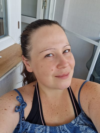 A woman with brown hair pulled back and a black bikini top with pink trim is shown in a close-up, slightly angled, outdoor selfie. She has light brown eyes and fair skin with numerous freckles and moles across her face, shoulders, and chest. She is smiling slightly and looking towards the camera. The background appears to be a balcony or patio area with a white railing and a wooden surface visible to the left. The scene is bright and suggests a warm, sunny day. This image was taken in Ovoshtnik, Bulgaria.