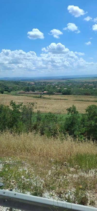 The image depicts a landscape view of a rural area, likely a village, under a partly cloudy blue sky. In the foreground, a metal guardrail is visible with gravel and sparse vegetation. The middle ground features fields of dry, golden grass and patches of green trees. Beyond that, a cluster of houses with red-tiled roofs, typical of Bulgarian architecture, are scattered across rolling green hills. The scene suggests a sunny day with a vast expanse of nature and human habitation. The metadata indicates the location is Kozar Belene, Bulgaria. There are no people or animals visible in the frame, nor any discernible actions or interactions. The overall environment is tranquil and bucolic.