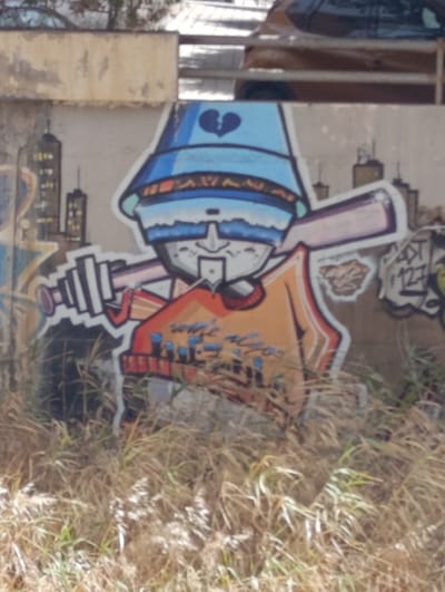 The image shows a graffiti mural on a concrete wall in Águilas, Spain. The mural depicts a cartoon character wearing a blue bucket hat with a broken heart on it, sunglasses, and an orange shirt. The character is holding a telescope-like object and appears to be looking through it. In the background of the mural, there are drawings of buildings, resembling a cityscape. The wall itself is weathered and stained, and the area in front of it is covered with dry grass and weeds. Above the wall, a portion of a car and a railing are visible. The graffiti style is bold and colorful, with clear outlines.