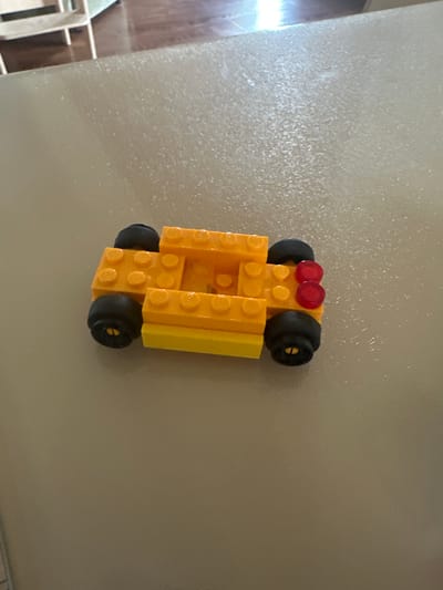 A small, orange and yellow Lego car is resting on a light-colored, textured surface. The car has four black wheels and two red taillights. The background shows a blurred wooden floor and what appears to be shelving. The image is taken from a top-down perspective. There is no visible indication of the city of Kiparisnoe, Ukraine.