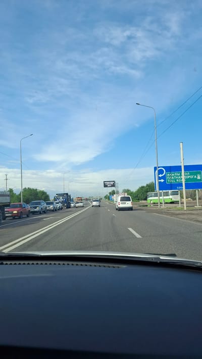 The image shows a highway scene in Панфилово, Kazakhstan. Traffic is heavy, with cars and trucks lined up on the road. A large blue sign indicating "Toll Road" is visible, along with overhead power lines and street lights. The sky is blue with scattered clouds.