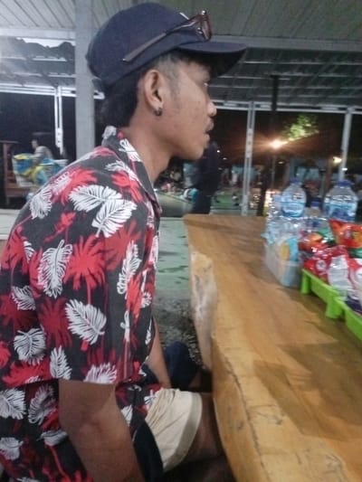 A young person, likely male, is seated at a rough-hewn wooden table or bench, viewed from a side profile looking towards the right of the frame. He wears a black cap with dark-rimmed sunglasses perched on top. His hair is dark, styled short on the sides with some length at the back, and he has a black hoop earring in his left ear. He is dressed in a short-sleeved, collared shirt featuring a pattern of red and white palm leaves against a black background, and light-colored shorts with a dark blue garment underneath.
On the wooden surface in front of him, several clear plastic bottles of water, identifiable by the brand "AQUA," are visible, along with an assortment of snacks packaged in colorful bags, arranged in light green plastic trays. The setting appears to be an open-air or semi-covered area, bustling with indistinct activity in the background. Artificial lights illuminate the scene, indicating it is nighttime. Structural elements like dark beams and a ceiling are visible above. The overall impression is that of a casual eatery or market stall in Pangalengan, Indonesia. The person's expression is neutral, and he seems to be observing something or someone outside the frame.