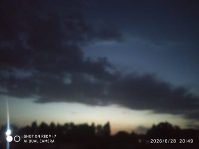 The image shows a dark, cloudy sky at dusk or dawn, with a faint band of lighter sky near the horizon. Silhouetted against the horizon is a line of trees. In the lower left corner of the image, there is a bright, blurred light source, likely a streetlamp or vehicle headlight. The text overlay in the lower left corner reads "SHOT ON REDMI 7 AI DUAL CAMERA" and in the lower right corner, "2026/6/28 20:49". The image appears to be a photograph taken outdoors in Khalkabad, Uzbekistan.
