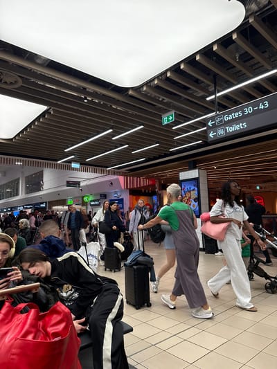 The image depicts a busy airport terminal, likely Luton Airport in the United Kingdom, filled with travelers. In the foreground, a young man with long dark hair is asleep on a bench, his head resting on his arm. A large red bag sits beside him. Behind him, other travelers are moving through the terminal, some with luggage. To the right, a woman in a grey jumpsuit and another woman in white athletic wear are walking. Above them, directional signs indicate "Gates 30-43" and "Toilets" with accompanying arrows and icons. Green exit signs are also visible on the ceiling. The terminal features modern architecture with suspended ceilings, exposed beams, and fluorescent lighting. Shops and other airport facilities are visible in the background. The overall scene conveys the typical atmosphere of an airport during a period of travel activity.