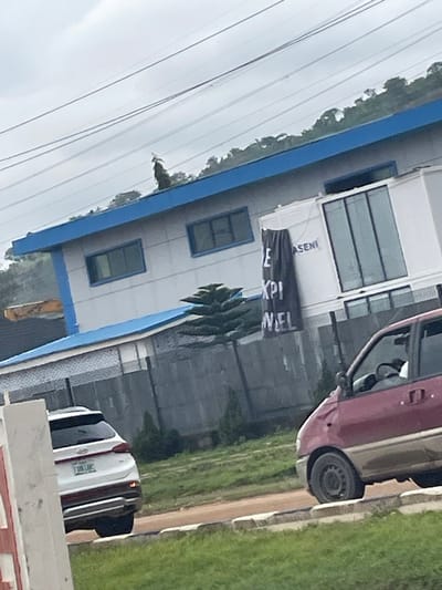 This image, taken in Jiru, Nigeria, depicts a street scene dominated by a multi-story building with a blue and white facade. The building has large windows and some signage, including a black banner with white text. A maroon car is parked to the right of the building, partially obscuring its side. In the foreground, several white vehicles are parked. The scene appears to be outdoors, with a grassy area visible to the right of the road, suggesting a suburban or commercial setting. The lighting suggests it is daytime, and the overcast sky hints at potentially cloudy or rainy weather. The image is oriented vertically, with the sky at the bottom and the ground at the top, as if the photographer was looking upwards at an angle.
Visible text on the black banner reads "E KPI NEL". The building also has text that appears to say "ASENI" above the banner.