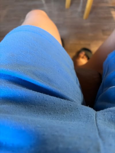 This media file shows a close-up view of a person's legs wearing blue shorts. The camera angle is from above, looking down at the subjects' legs and the fabric of the shorts. The fabric of the shorts appears to be a light blue and has a textured surface with small raised dots, possibly from the material itself or pilling. The person is wearing dark-colored sandals. In the background, a wooden floor is visible, along with what looks like parts of chairs or furniture. The lighting suggests an indoor setting. No text is visible in the image. The image is taken from a low perspective, emphasizing the texture and color of the fabric and the casual attire of the individual.