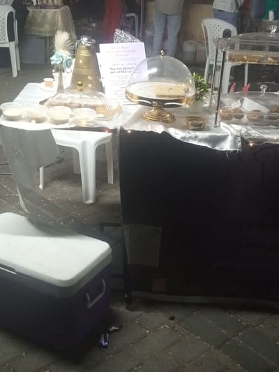 The scene depicts an outdoor market stall in the Makadara/Mji Wa Kale ward, Kenya, likely during evening hours. In the foreground, a dark blue cooler box with a white lid rests on interlocking grey pavers. A retail display table, covered with a white tablecloth over its top and a dark fabric skirt below, dominates the mid-ground. Small, warm-toned string lights are visible along the table's edge.
On the table, from left to right, are: four small, clear plastic containers with white lids, each holding a light-colored substance; a small white object, possibly a cup; a tall, golden, traditional-style carafe with a lid; a clear vase containing a white feather and light blue artificial flowers; a white sign advertising "MohaSweet at the Bazaar!" with "SPECIAL OFFERS"; a golden pedestal cake stand with a clear dome cover, displaying a rectangular dessert; a small golden rectangular box; and a two-tiered clear plastic display case containing multiple small pastries. A small pot with green artificial foliage is situated between the cake stand and the tiered display. A white plastic stool supports the table from beneath, and another white plastic stool/chair is partially visible in the blurred background.
In the background, several blurred figures of individuals are discernible, wearing casual attire such as jeans. Other market stalls with draped tables and white plastic chairs are also present. The ground is paved with dark grey interlocking bricks. A small piece of blue and silver refuse lies on the ground near the cooler box. No direct human interactions are clearly depicted in the foreground.