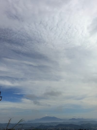 An expansive sky filled with white, textured clouds dominates the image. Below, a hilly landscape stretches into the distance. The terrain consists of forested areas and possibly small towns, fading into a misty blue with a prominent mountain range. Some grass is present in the foreground, and a metallic object is visible in the bottom-left corner. The overall impression is one of a vast, open vista.