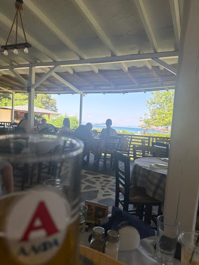 This image depicts a daytime scene at an open-air restaurant or tavern located in Ολυμπιάδα, Greece, overlooking a beach and the Aegean Sea. The foreground is dominated by a blurred, out-of-focus glass or bottle, exhibiting a white circular label with a prominent red capital 'A' and blue Greek text (ΑΛΦΑ) below it.
The restaurant features a rustic architectural style with a wooden ceiling supported by white-painted wooden beams and posts. A light fixture, comprising a dark wooden bar with three exposed bulbs suspended by thick ropes, hangs from the ceiling. The floor is an irregular mosaic of light-colored stones.
On a table in the lower right, covered with a white tablecloth, are two silver shakers (salt and pepper), a white spherical object, a folded blue napkin, and an empty clear glass. A small menu or sign with a QR code is visible behind these items.
Several patrons are seated at dark wooden tables with checkered blue and white tablecloths. Most individuals are seen from the back or side, with details obscured by distance and focus. One person on the mid-right is identifiable by a dark shirt and glasses.
Beyond the restaurant's railing, a bright blue sea with distant landforms stretches to the horizon under a clear blue sky. A narrow strip of beach is visible, populated by several straw beach umbrellas. Lush green trees and vegetation line the coastline, indicating a natural environment.
