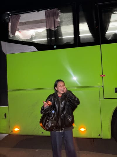 The scene, set at night in Hattencourt, France, features a lime green VDL Futura coach bus parked on a paved surface. The bus exhibits tinted windows, some revealing rumpled interior curtains, and an illuminated green emergency exit pictogram on the forward upper left window. Multiple amber side marker lights are active along its lower panels, with a bright light reflecting off its side. A single individual stands center-right in the foreground, captured in motion, laughing with their right hand covering their mouth. They are dressed in a dark bomber jacket over a dark shirt, dark wide-leg pants, and carry a dark shoulder bag with an attached clear plastic bottle. A copper-colored disposable cup is held in their left hand.