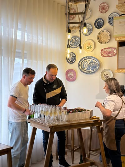 The image portrays an indoor scene within a modern establishment in Felgueiras, Portugal, set up for a social gathering or tasting event. Three individuals are positioned around a rectangular wooden table.
To the left, a male subject in a white short-sleeved t-shirt and light blue jeans is opening a dark green bottle over the table. In the center, a male subject, identified by a black chef's jacket with a beige diagonal stripe and a "Chocolate" logo, stands looking down at the table's contents. To the right, a female subject with shoulder-length grey-blonde hair, wearing a white short-sleeved blouse, dark jeans, and a brown apron, holds a small metallic plate with food.
The table is laden with multiple clear glass champagne flutes, most filled with a pale yellow liquid. Adjacent to the glasses is a rectangular white dessert, likely a cake, topped with an arrangement of red berries, blueberries, and green leaves. A small dish with a pastry or snack is visible in front of the female subject. A green bottle rests on the floor beneath the table.
The background features a large window on the left, covered by floor-length translucent white vertical blinds. The white wall is decorated with an array of antique-style decorative plates of various sizes and patterns, predominantly in blue and white, red, and pink. Above the plates, a chandelier-like fixture constructed from a wooden ladder-like frame suspends multiple exposed filament light bulbs. On the right, a light-colored wooden shelving unit displays packaged goods, with visible text including "SÃO LUIZ Winemaker" and "Quinta Nova."