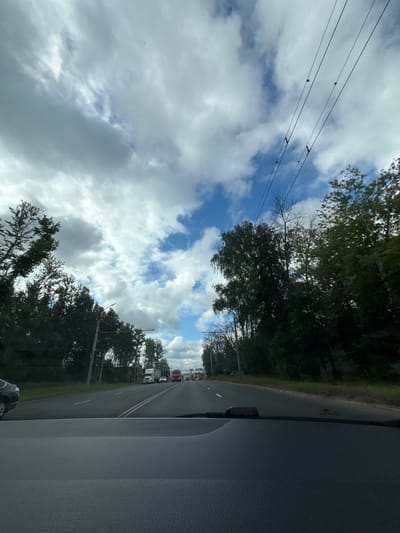 The image captures a view from inside a vehicle on a road in Korobovo, Russia, on a partly cloudy day. The road ahead is paved with asphalt, with a double yellow line separating lanes of traffic. Several vehicles, including trucks and cars, are visible on the road, moving in the same direction. On either side of the road, there are dense trees lining the area. Overhead, power lines with visible insulators are strung across the sky, which is a mix of blue sky and white and gray clouds. The foreground of the image is dominated by the dashboard of the vehicle from which the photo is taken.