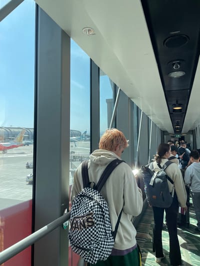 The image captures the interior of an airport jet bridge at Muscat International Airport in Seeb, Oman, under clear, sunny skies. A queue of passengers proceeds along the enclosed walkway, which features large, floor-to-ceiling windows on the left side. Through these windows, an airport tarmac is visible, displaying a red and white aircraft, other distant aircraft tails, and white ground support vehicles. The jet bridge's ceiling is white, with a central black panel housing multiple recessed lights and ventilation grilles. A metal handrail extends along the base of the windows. In the immediate foreground, from a rear perspective, a person with reddish-blonde hair wears a light beige hooded sweatshirt, dark green shorts with a yellow stripe, and a black and white checkered Vans backpack. Directly behind this individual, another passenger, also viewed from the back, wears a light-colored ribbed long-sleeved top, dark pants, and carries a dark grey backpack. A bright reflection appears on the back of this second passenger. Further down the jet bridge, more passengers are visible in the queue, uniformly moving towards the aircraft boarding point.