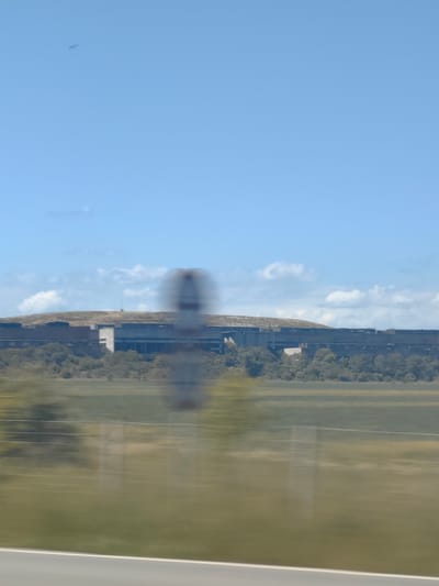 The image depicts a landscape in Shishmantsi, Bulgaria, with a long, low building situated on a plain against a backdrop of a hill partially covered in vegetation under a partly cloudy sky. The building appears to be large and potentially industrial or institutional. A layer of trees separates the building from the foreground. A blurred sign is visible in the foreground. A road is at the very bottom of the view. The overall scene appears to be taken from a moving vehicle, indicated by the motion blur.