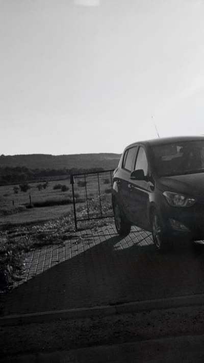 The image shows a black and white view of a dark colored car parked on a paved surface. The car is positioned on the right side of the frame, with its front end facing towards the viewer and slightly to the left. The car's headlights and part of its grille are visible. Behind the car, a black metal fence runs horizontally across the mid-ground. Beyond the fence, there is a field or pasture with some scattered vegetation. In the distance, rolling hills covered in trees form the horizon. The sky is bright and overcast. The paved surface in the foreground is made of rectangular bricks laid in a pattern, and it casts a distinct shadow from the car and fence. The scene appears to be outdoors, possibly in a rural or semi-rural area. The location is identified as Horesti, Moldova. There are no people visible in the image.