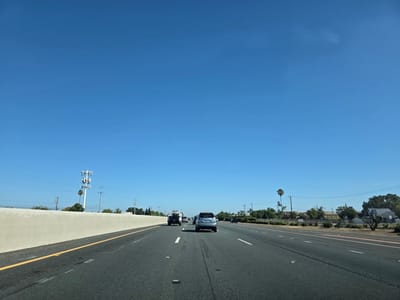 The image depicts a highway scene in Elk Grove, California, United States of America. The perspective is from inside a moving vehicle, looking forward. The sky is clear and blue. The highway has multiple lanes of asphalt with white lane markings and reflectors. A concrete barrier runs along the left side of the highway. On the left, beyond the barrier, a cell phone tower is visible. On the right side of the highway, there are trees, power lines, and residential buildings, including a house with a "45" speed limit sign. Several vehicles are on the highway, including a silver SUV in the foreground, a white pickup truck towing a trailer behind it, and other cars further ahead. The overall environment suggests a typical suburban highway setting on a sunny day.