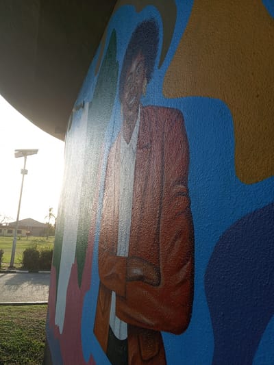 A mural depicting a person with dark hair wearing an orange suit, a white shirt, and earrings is painted on a wall. The mural's background features broad strokes of blue and brown. The wall is located outdoors in a grassy area, likely in Tawpo, Nigeria, as specified. A road and a streetlamp are visible in the background along with several buildings. The light suggests a sunny day or early morning. The mural appears to be on a structure, possibly a building or overpass.