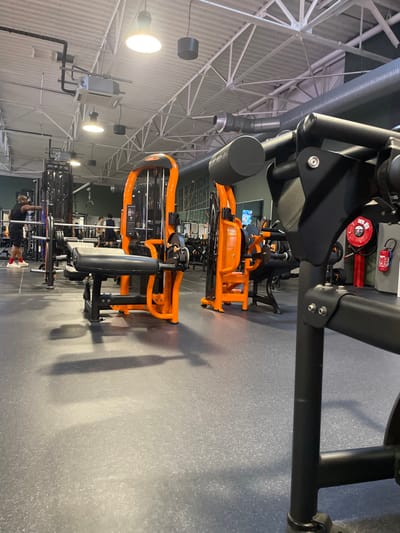 The image depicts the interior of a Basic-Fit gym located at 15 Rue de l'Institut, 14200 Hérouville-Saint-Clair, France. The scene captures a wide view of an exercise area with a dark gray, speckled rubberized floor and an industrial-style ceiling featuring white metal trusses, exposed silver and black ventilation ductwork, and multiple large pendant light fixtures.
In the immediate foreground, a black exercise machine frame with a black padded roller is visible on the right. Centrally positioned is an orange and black leg extension/curl machine with black padded seating. To the left, a male subject, dressed in a black sleeveless shirt, black shorts, and red athletic shoes, stands facing a black barbell rack equipped with a silver barbell. His left arm is extended forward, seemingly in preparation for or completion of an exercise. In the mid-ground and background, several other individuals are present, engaged with various pieces of orange and black exercise equipment, including cable machines and weight benches. The rear wall is a light green/teal color, bearing partial white lettering. On the far right wall, a red fire extinguisher and a red fire hose reel are mounted. The overall environment is brightly illuminated by overhead lighting, casting shadows on the floor.