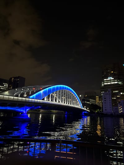 A nighttime, medium-shot, eye-level, vertical view captures the Kiyosu Bridge, a prominent landmark in Koto, Japan. The bridge, illuminated with vibrant blue lights, spans across a dark body of water, likely the Sumida River. The architectural structure of the bridge is clearly visible, with its arched superstructure glowing intensely. The reflections of the bridge's blue lights and the lights from surrounding buildings shimmer on the water's surface, creating a dynamic visual effect. In the background, a cluster of modern high-rise buildings with illuminated windows forms the city skyline. The sky above is dark and cloudy, with only a faint hint of diffused light. The scene is devoid of any visible people or animals, focusing solely on the urban landscape and the illuminated bridge. A dark railing is visible in the foreground, suggesting the vantage point is from a nearby walkway or embankment. The overall atmosphere is one of urban nighttime beauty and tranquility.