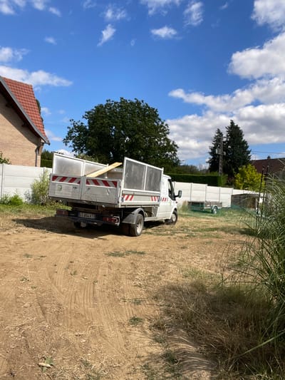 In the city of Viry-Noureuil, France, on a sunny day with scattered clouds, a white flatbed truck is parked on a dirt lot. The truck bed is filled with a wooden plank and is backed by a silver metal enclosure with red and white reflective stripes. A white concrete fence runs behind the truck, separating the lot from a grassy area with a small trailer. A house with a red-tiled roof is visible to the left of the truck. Trees and vegetation surround the scene, with tall grass in the foreground. The license plate of the truck reads "CT 854 XT".