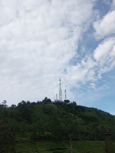 A lush green hill covers the lower portion of the image. Terraced rows resembling tea plantations span across its base. The hill is densely covered with trees. Atop the hill, several communications towers rise against a cloudy sky with patches of blue visible. Power lines are visible in the lower right corner, with poles holding them.
