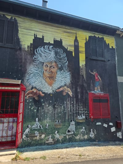 The mural depicts a fantastical scene on the exterior wall of a building, possibly in the city of Viterbo, Italy, though this cannot be confirmed solely from the image. A large, imposing elderly woman with wild, white hair and a fur collar dominates the central portion of the mural. Her expression is a mischievous grin, and her hands are outstretched as if casting a spell. She is positioned against a dark, almost misty background with faint, glowing orbs, suggesting an enchanted or magical setting.
In the upper section of the mural, a stylized silhouette of a city skyline is painted, featuring buildings that resemble London landmarks like Big Ben and possibly the London Eye. This urban backdrop is rendered in dark grays and yellow hues, creating a dramatic contrast with the foreground.
To the left of the main figure, a classic red British telephone booth is depicted, its door ajar, revealing floral patterned curtains. To the right, a red post box is mounted on the wall, with a small depiction of a young boy in a red shirt and blue shorts standing atop it, reaching towards the city skyline.
The lower portion of the mural features a grassy landscape populated by numerous Dalmatian puppies playing in baskets and in the grass. Their black and white spots stand out against the green. The wall itself appears weathered in some areas, with peeling paint and exposed brickwork visible beneath the mural. A few scattered envelopes are painted on the wall to the right of the post box. The scene is illuminated by bright daylight, casting shadows on the ground in front of the wall.