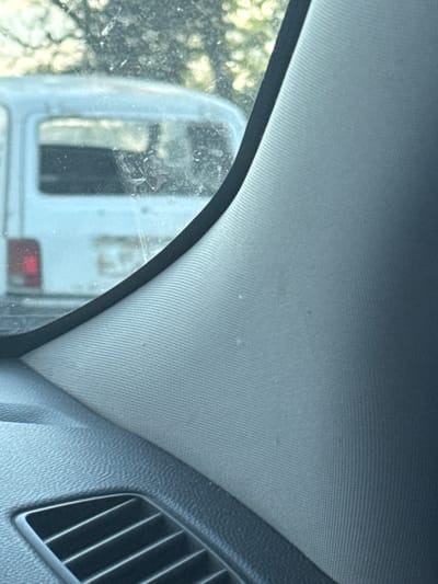 The image shows a view from inside a vehicle, looking out the side window. The window appears to be dirty, with smudges and streaks. Outside the window, another vehicle, possibly a light-colored van, is visible. The interior of the car features a dark-colored dashboard with a visible air vent in the lower left corner. The background outside the vehicle is blurred, suggesting foliage and trees. There are no people or discernible activities depicted in the scene. The image does not contain any information to place it in Gaevka, Russia.