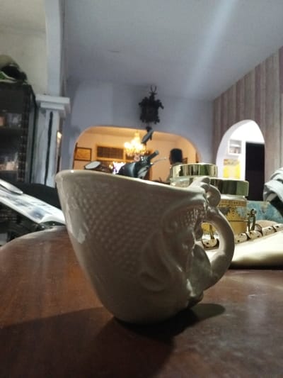 This image depicts an indoor scene in the city of Longmiang, Laos. In the foreground, a light-colored, possibly ceramic, mug with a textured, scale-like pattern and an ornate handle rests on a dark wooden surface. A shadow from the mug extends across the table. Behind the mug, to the right, several small, clear jars with gold-colored lids are visible. To the left, an open magazine or book lies on a dark surface. The midground shows a room with light-colored walls and ceiling. A dark, ornately carved cuckoo clock hangs on the wall. Two arched doorways or openings lead to other parts of the interior. Through the left arch, a warm-toned chandelier is visible, illuminating the background, and the silhouette of a person, facing away, is present. A partial view of a dark scooter or motorcycle's handlebars and mirror is visible below the cuckoo clock, within the left arched area. The right arched opening reveals a room with light-colored vertical striped wallpaper and a further doorway. The overall lighting is subdued. No direct actions or interactions are depicted.