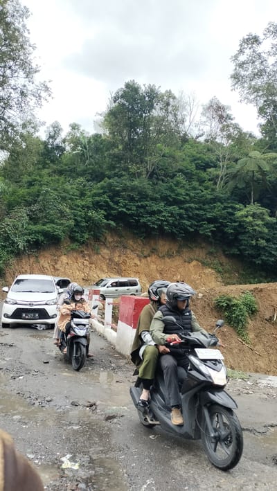In Bintangpermata, Indonesia, a cloudy day scene unfolds on a wet, pothole-ridden road that winds uphill through a lush, forested landscape. A white car, with license plate BL 1423 JM, is parked on the left side of the road. Further up the road, a silver minivan is visible. Two motorcycles, each carrying passengers, are moving up the incline. The motorcycle in the foreground has two riders, a driver wearing a black helmet and a dark vest over a green jacket, and a passenger in a khaki-colored cloak, with their face obscured by a helmet. The motorcycle behind it has a driver wearing a white helmet and a passenger with their face obscured by a helmet, both wearing protective gear. The road surface is dark and appears muddy and uneven, with visible water accumulation in the depressions. Red and white barriers line the side of the road. The background is dominated by dense green foliage covering the hillside, with a few taller trees and ferns interspersed.
