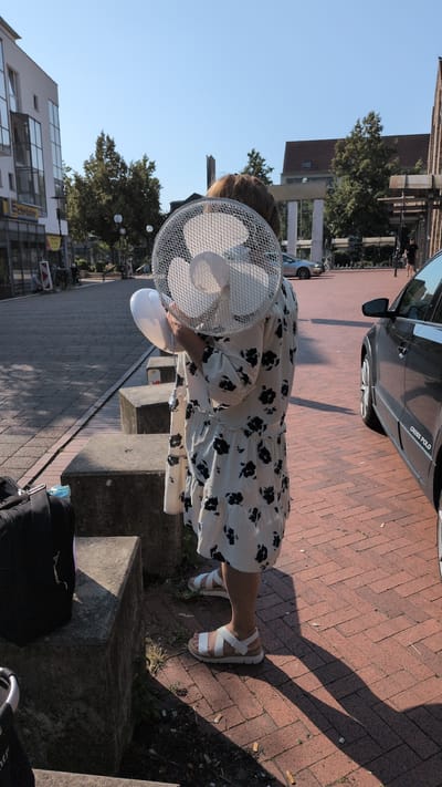 A person is standing on a paved sidewalk next to a street in Osnabrück, Germany, holding a portable electric fan in front of their face. The person is wearing a light-colored dress with a black floral pattern and white sandals. The fan is silver and white. To the right of the person, a black car with "CROSS POLO" written on its side is parked. In the background, there are buildings, trees, and a clear blue sky. The scene appears to be a public outdoor space, likely a street or plaza.