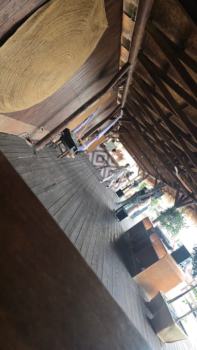 The image depicts an outdoor venue with a wooden deck and a thatched roof. Several people are present, some of whom appear to be staff or performers, wearing light-colored outfits. There's a DJ setup with turntables and speakers, suggesting a musical event is either about to happen or is in progress.
The scene has a relaxed, possibly festive atmosphere. The lighting suggests it is daytime, likely bright given the shadows. In the background, there are planters with greenery, and what looks like a building or structure. The overall setting appears to be a resort, beach club, or an outdoor entertainment space.
Notable details include the woven rug on the left side of the deck, the patterned design on what could be a bar or counter in the middle ground, and various pieces of audio equipment. The perspective is slightly tilted, giving a dynamic feel to the image.
No visible text is present in the image. The location context provided is Parakar, Armenia.