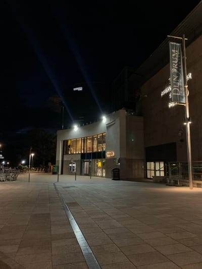 The image depicts the Dun Drum Town Centre located in Dublin 16, Ireland, at night. A street sign labeled "Dun Drum Town Centre" is illuminated above the entrance of the center. A brick-paved walkway is beside the building. The building appears to house establishments such as McDonald's and a restaurant named "Penthouse." The sky is dark, and streetlights provide illumination. A line of bicycles are parked close to the street.