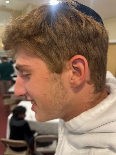 This image depicts a young male in an indoor setting within Bergenfield, United States. The primary subject, positioned in the right foreground and mid-ground, is shown in profile, facing left. He has light brown, wavy hair and wears a dark kippah partially visible on the top of his head. He exhibits light stubble on his cheeks and jawline, a prominent nose, and a small red mark on his right cheek. He is wearing a white hooded sweatshirt, with the hood's drawstrings visible at the neck. His mouth is slightly open.
The background is out of focus, showing multiple individuals and environmental elements. One person wearing a dark green shirt is visible standing in the distant left. Another individual with dark hair, dressed in a dark top, is seated at a table covered with a white tablecloth in the mid-ground left. The floor is covered with a red carpet. Light-colored folding chairs are discernible around tables. The upper background features light-colored walls, and bright light sources create lens flares, notably above the primary subject's head. The scene suggests a communal gathering or event in a hall.