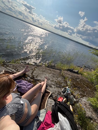 The image depicts two individuals relaxing on a rocky shore overlooking a wide body of water under a bright, cloudy sky. The water reflects the sunlight, creating a shimmering effect. On the rocks, there are several objects, including a black backpack, a white plastic bag, some clothing items (a pink garment, a purple towel, and a striped top), several empty cans, and a pair of shoes. One person is visible from the back, wearing a striped top, with their legs extended. The feet of another person are also visible, positioned near the water's edge. The environment suggests a casual outdoor setting, possibly a park or a recreational area near a lake or river. The presence of cans and a backpack implies that the individuals have been picnicking or spending leisure time outdoors. The location is identified as Shuiskaya Chupa, Russia.