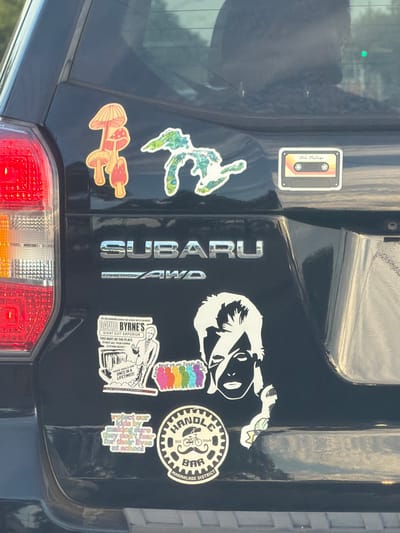 The image captures the rear tailgate of a dark-colored Subaru vehicle, likely an SUV or hatchback, adorned with numerous bumper stickers. The car's "SUBARU" and "SYMMETRICAL AWD" emblems are visible in the center. A portion of a red taillight is seen at the bottom.
On the left side of the tailgate, there is a vintage-style cassette tape sticker labeled "Utah Mixtape," a green and blue abstract sticker resembling a frog or gecko, and a cluster of orange and red mushroom stickers.
On the right side, a black and white stylized decal of a person's head or bust is prominent. Below it, a rainbow-colored sticker features human figures and the text "EVERY BODY IS AN IDEAL BODY." To its right is a circular sticker with a bicycle logo, identifying it as "HANDLE BAR," "SLC," "Utah," and "MARMALADE DISTRICT." Further down, a rectangular sticker advertises "DAVID BYRNE'S GIANT SUIT EMPORIUM," with text including "THIS MUST BE THE PLACE," "TO MEET ALL YOUR FORMAL CLOTHING NEEDS!," and "THESE DEALS ARE ONCE IN A LIFETIME!!!" It also depicts a skeleton in a suit. Below this, a text-only sticker reads: "protect our kids by making sure they don't fear for their lives at school."
The image appears to be taken during the daytime, with reflections on the car's surface suggesting bright lighting. No specific activity beyond showcasing the personalized vehicle is evident. The general location context is Arlington Heights, United States of America.