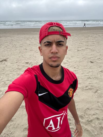A young man takes a selfie on a sandy beach. He wears a red cap and a Manchester United soccer jersey. The background shows a wide beach meeting the ocean under an overcast sky. In the distance, a single figure can be seen walking along the shoreline. The image is taken at an angle, showing both the man and the expansive beach setting.